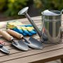 A neatly arranged collection of essential home gardening equipment including trowels, pruning shears, and a watering can placed on a wooden garden table