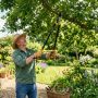 Gardener using long-handled bypass loppers to prune a tree branch in a sunny backyard garden